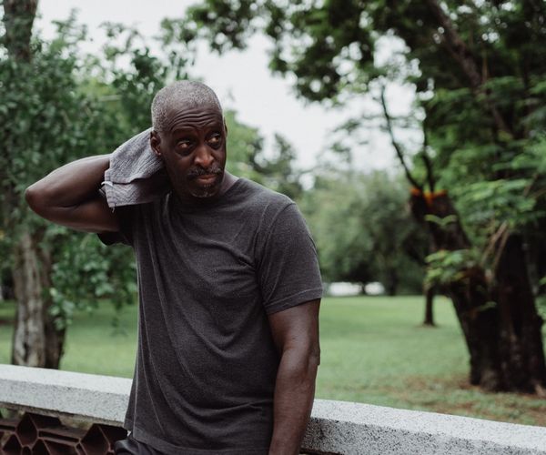 A man standing in a park, drying his neck with a towel after a run 