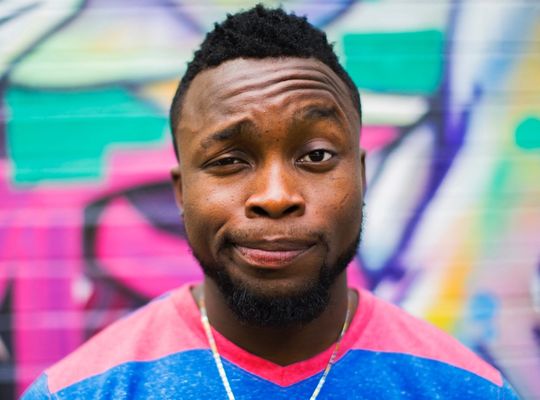 A man smiling with his eyebrow raised standing in front of a colorful graffiti background